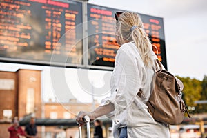 Woman passenger with backpack is checking arrival and departure board at railway station