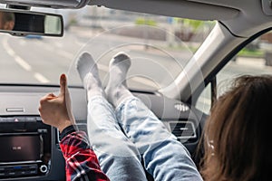 Woman passenger, legs in a jeans and socks on car dashboard while travel on the highway.  Girl showing thumbs up . Freedom