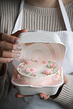 Woman pastry shef wearing white apron and holding small bento cake decorated with cream cheese pink flowers. Woman hands hold