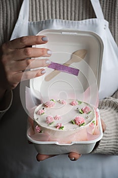 Woman pastry shef wearing white apron and holding small bento cake decorated with cream cheese pink flowers. Woman hands hold