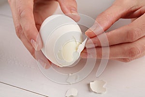 Woman peeling boiled egg at white wooden table, closeup