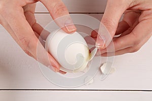 Woman peeling boiled egg at white wooden table, top view