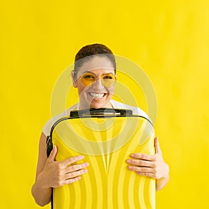 A woman with a perfect snow-white smile in sunglasses hugs a suitcase on a yellow background. Excited girl waiting for