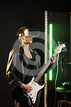 Woman performer playing at electric guitar in sound studio while wearing vr goggles for concert simulation