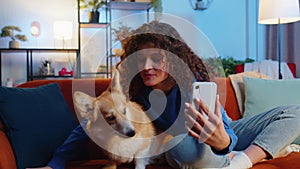 Woman pet lover kissing cute corgi dog and taking selfie via smartphone on sofa at home living room