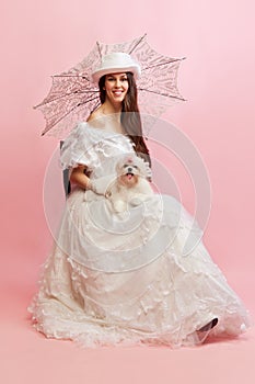 Woman and pet. Smiling. Portrait of beautiful lady in white vintage dress posing with dog over pink background. Concept