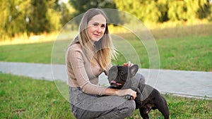 Woman pets a French bulldog, crouching next to him during a walk in the park on a summer day, looking at him lovingly