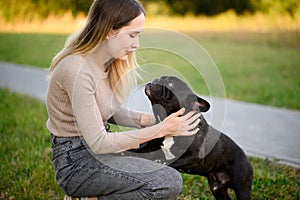 Woman pets a French bulldog, crouching next to him during a walk in the park on a summer day, looking at him lovingly