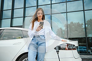 Woman with phone near an rental electric car. Vehicle charged at the charging station