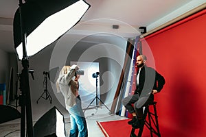 Woman photographer making business portraits for handsome bearded man on red background in photo studio. Work of a photographer.