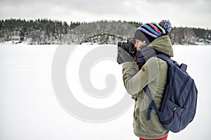 A woman photographer in warm clothes and a backpack is holding a camera in her hands and taking pictures of a winter landscape.
