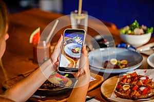 Woman photographing dinner in restaurant through mobile phone at table. Hand hold smartphone taking photo of dish before