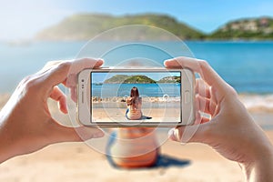 Woman photographing other girl sitting in the beach. She is using to take a photo the camera of a smartphone.