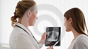 Woman physician showing X-ray on tablet to female patient in clinic. Female professional doctor at work