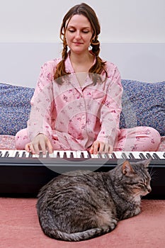 Woman pianist in red pajamas playing electric piano on home bed next to cat