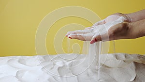 Woman picks up palms full of white dry sand on yellow studio background. Sandy particles wake up through your fingers
