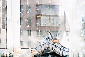 Woman in pijamas sitting on windowsill with a cup of coffe and phone. Winter mood