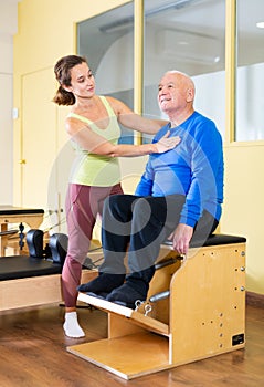 Woman pilates instructor helping senior man exercising on combo chair