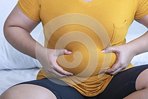 Woman pinches fat on her belly large size while sitting on white bed at home. Upset female suffering from extra