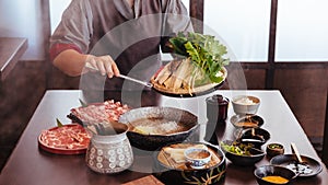 A woman pinching vegetables into hot pot by tongs with Wagyu A5 beef and sliced Kurobuta in Shabu clear soup with stream