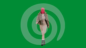 Woman in pink balaclava, tiger fur coat and evening dress walking. Woman freak on green background in studio. Fashion