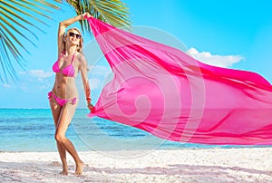Woman in pink bikini holding pink fabric in wind on the tropical beach