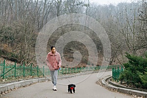 Woman in pink coat walks down the alley in the park with a funny little dog in a sweater in the cool spring morning, leisure on a