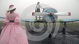 A woman in a pink dress of 60th comes to a vintage airplane on which stands the pilot