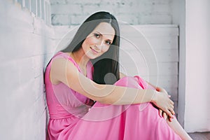 Woman in a pink dress sits leaning on the wooden white wall. He looks at the camera and smiling