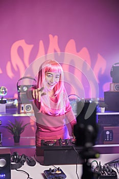 Woman with pink hair playing techno song at professional turntables while filimg process with mobile phone camera