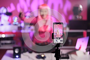 Woman with pink hair playing techno song at professional turntables while filming music process with phone camera