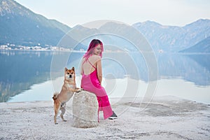 A woman with pink hair shares a lakeside moment with a Shiba Inu dog