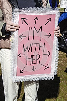 Woman in pink jacket hold protest pink sign edged with lace - Im with her - with arrows in every direction - solidarity - feminism