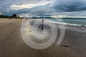 Woman with pink leggings and wide brimmed hat walking along wet beach under stormy skies in Byron Bay Australia with other beach