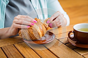 Woman with pink manicure is tearing a small piece from large croissant