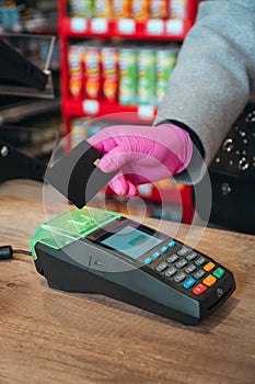 Woman in pink protective glove paying with contactless card at food store during quarantine. Closeup view of terminal