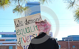 Woman n pink hat with back to camera stands against city skyline with sign We Demand Equality Diversity at Womens