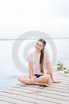 A woman in a pink t-shirt sitting in a wooden pier front of water.Brown hair,smile
