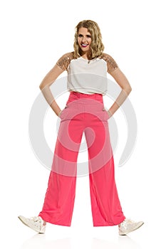 Woman In Pink Wide Legs Trousers Is Standing Legs Apart, Holding Hands In Pockets And Smiling