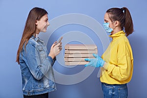 Woman from pizza delivery service wearing yellow shirt, protective mask and gloves giving food order and holding pizza boxes