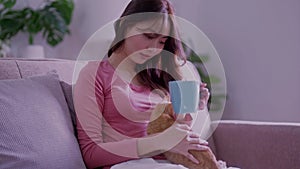 woman in a plaid drinking hot tea, petting a relaxed cat while stroking a cat sitting on her lap on the sofa at home