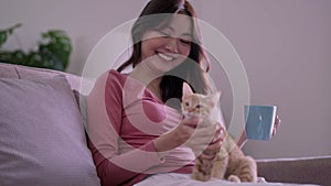 woman in a plaid drinking hot tea, petting a relaxed cat while stroking a cat sitting on her lap on the sofa at home