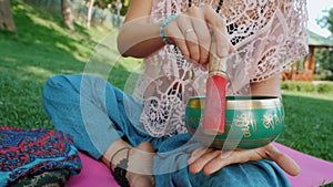 Woman playing singing bowl while sitting on pink yoga mat in park at summer. Vintage tonned. Beautiful girl with mala