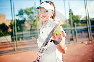 Woman playing tennis, holding racket and ball. Attractive brunette girl wearing white t-shirt and cap on tennis court