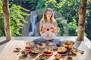 Woman playing on Tibetan singing bowl while sitting on yoga mat against a waterfall. Vintage tonned. Beautiful girl with