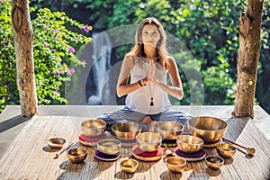Woman playing on Tibetan singing bowl while sitting on yoga mat against a waterfall. Vintage tonned. Beautiful girl with