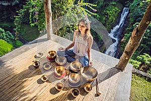 Woman playing on Tibetan singing bowl while sitting on yoga mat against a waterfall. Vintage tonned. Beautiful girl with