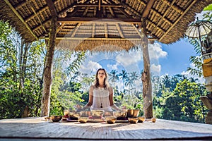 Woman playing on Tibetan singing bowl while sitting on yoga mat against a waterfall. Vintage tonned. Beautiful girl with