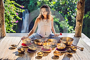 Woman playing on Tibetan singing bowl while sitting on yoga mat against a waterfall. Vintage tonned. Beautiful girl with