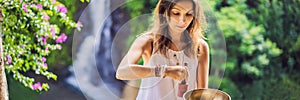 Woman playing on Tibetan singing bowl while sitting on yoga mat against a waterfall. Vintage tonned. Beautiful girl with